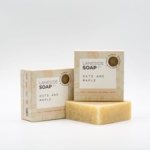 A picture of a Laneside Soap Company oats and maple soap bar sat slight in front of a packaged version of the product, with another packaged version of the product sat on top of it.