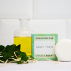 A photo of a Laneside Soap Company minty fresh shampoo bar leant against it's packaging, alongside some essential oil in a glass bottle and some decorative greenery