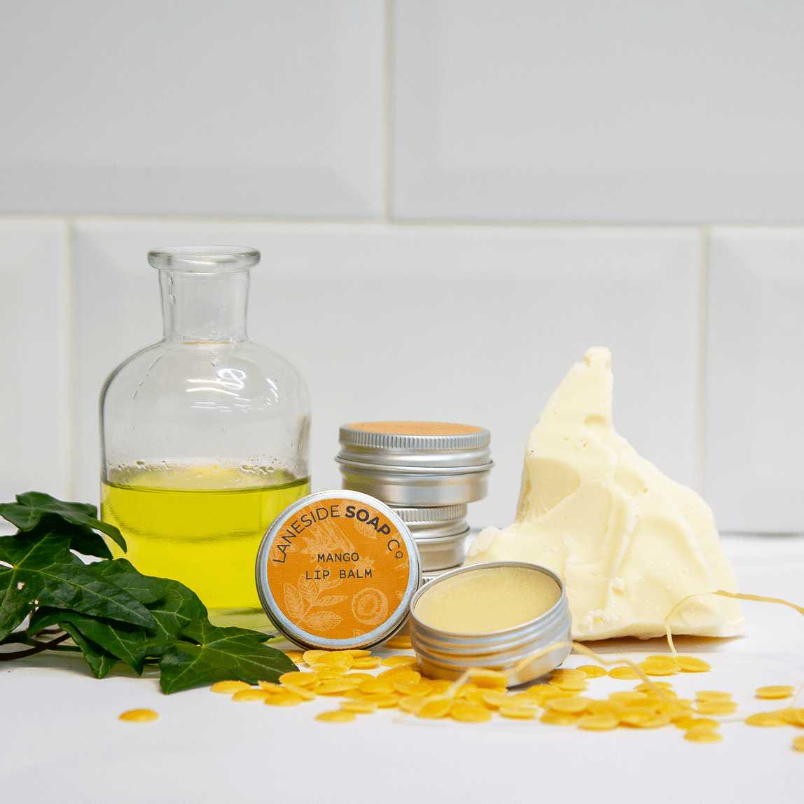 A photo of an open tin of Laneside Soap Company mango lip balm sat in front of some closed tins, stacked on top of each other alongside some essential oil, a piece of mango and some greenery