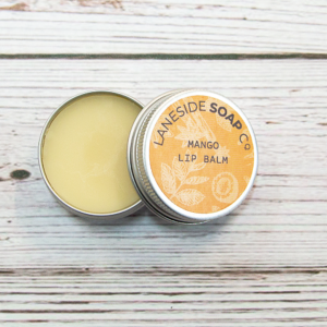 A tin of Laneside Soap Company Mango Lip Balm, with the lid opened and leaning on the side of the tin, revealing the lip balm inside.